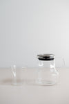 KINTO Cast glass teapot with stainless steel lid beside glass tumbler from the side view.