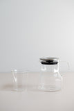 KINTO Cast glass teapot with stainless steel lid beside glass tumbler from the side view.