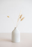 Ceramic bud vase with narrow mouth in white with natural specs from the side view holding a few dried florals.