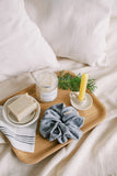 An 6 inch natural tube beeswax candlesticks made with pure Canadian beeswax in a teacup candle holder with a Holiday spa gift set that includes a natural bar soap, bath soak, ceramic soap dish, linen scrunchie and Korean exfoliating towel set on a wooden tray with pine leaves on top of a bed.